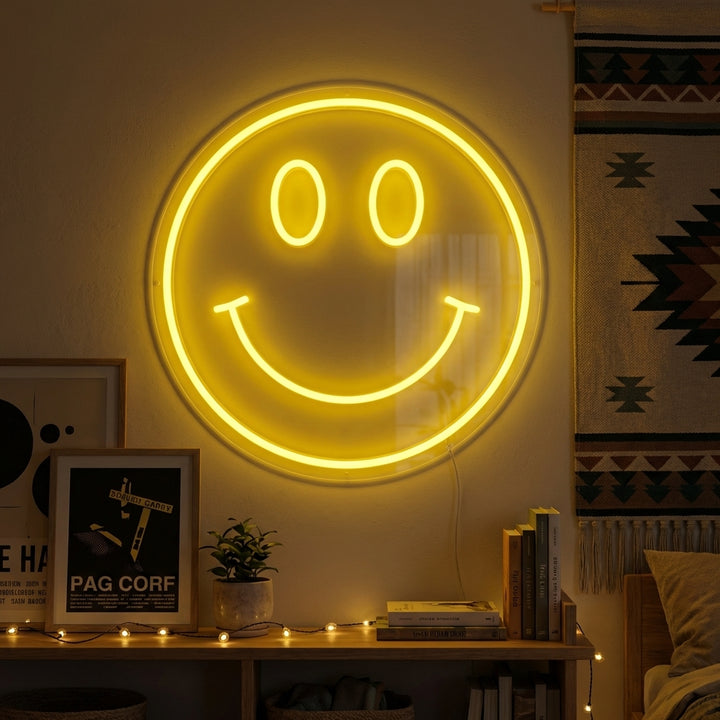 A bright yellow 'Element Symbol Emoji Smile Face' neon sign glowing on a textured bedroom wall, surrounded by pop art posters and fairy lights.