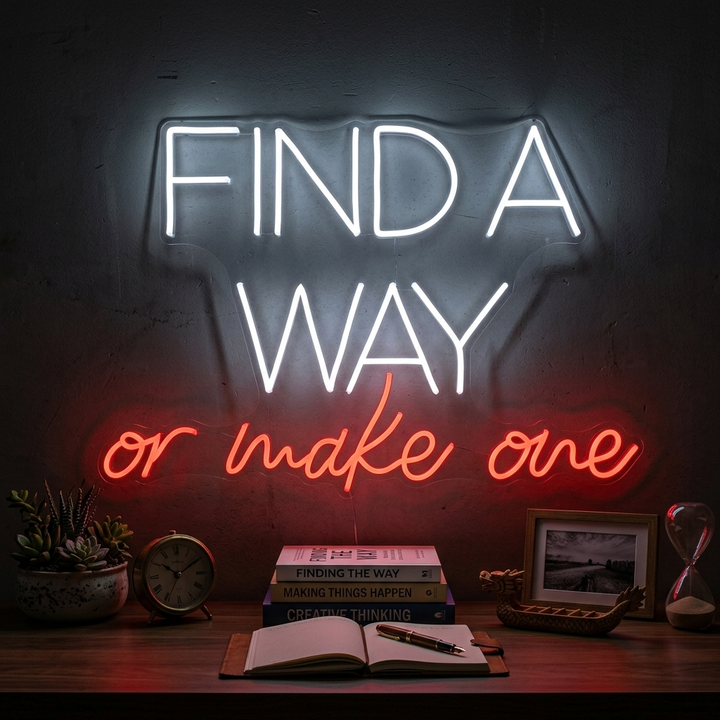 A bright pink 'Find A Way Or Make One' neon sign glows on a textured gray wall above a stack of motivational books in a modern home office setting.