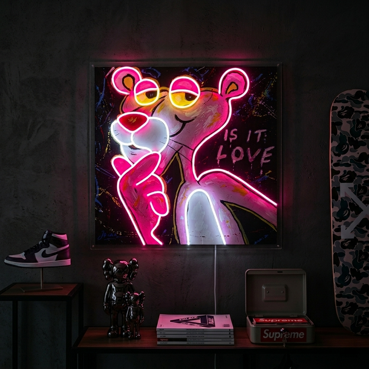 A physical photograph showing the "Pink Think Panther, Is It Love" UV print neon sign glowing on a textured bedroom wall above a console table.