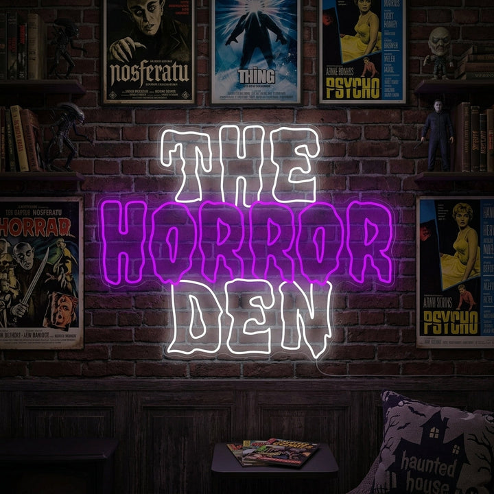 "The Horror Den" neon sign glowing on a wall in a moody, dark room with a plush armchair and spooky decorations.