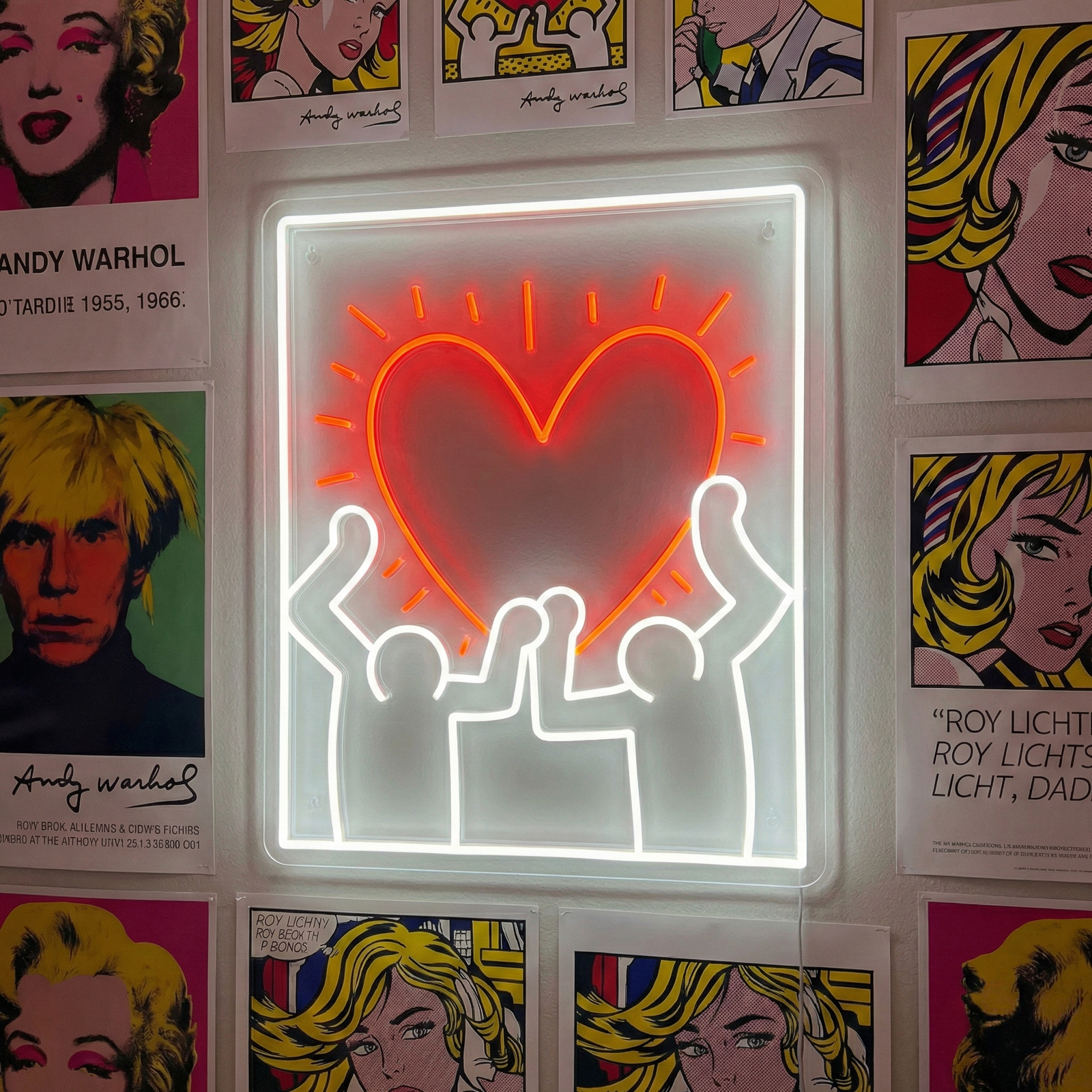 A vibrant 'Radiant Heart' neon sign with a glowing outline and solid heart center, hanging on a brick wall alongside artistic posters and pop art. A person is seated below looking towards the sign.