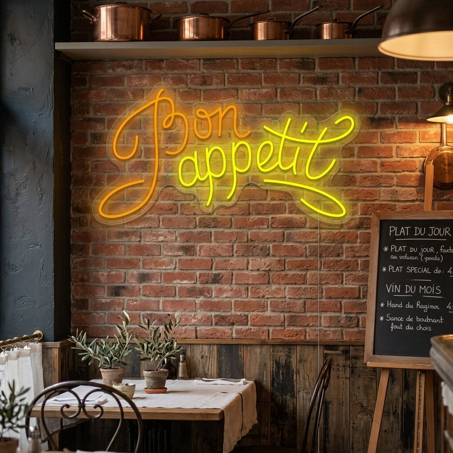 A glowing 'Bon Appetit' neon sign in a stylized script, mounted on a rustic wooden plank wall above a cozy home kitchen counter with dining accessories in a warm room setting.