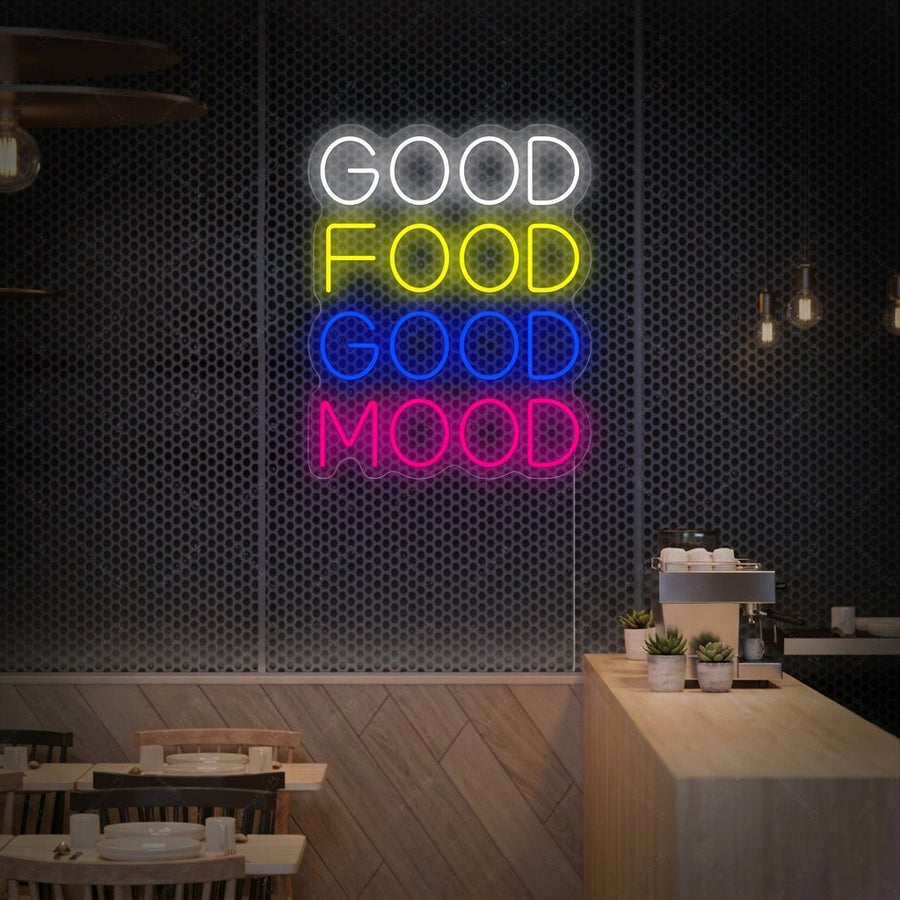A vibrant 'Good Food Good Mood' neon sign in script font glowing pink on a textured white kitchen wall above a wooden countertop with plants and kitchenware.