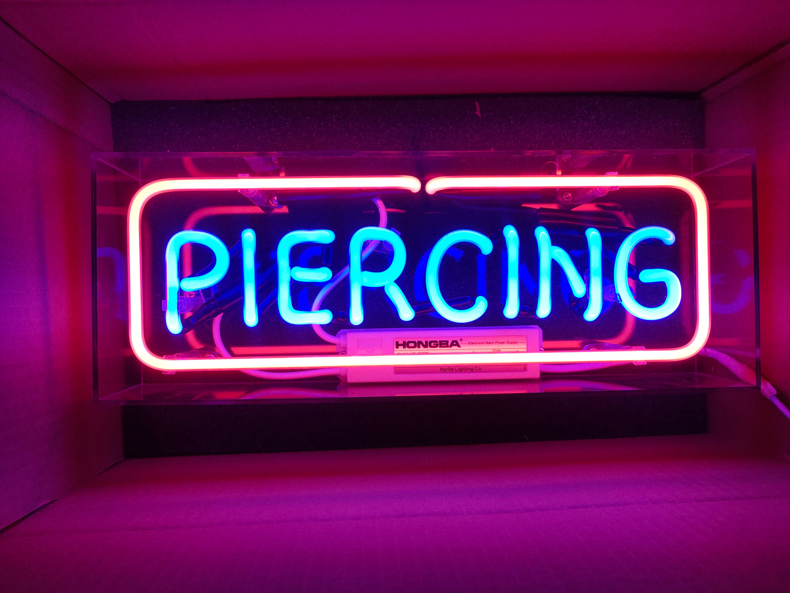 A 'Piercing' acrylic box neon sign with pink and blue glass tubing is shown inside its packaging box, revealing manufacturing details like the power supply and mounting hardware.