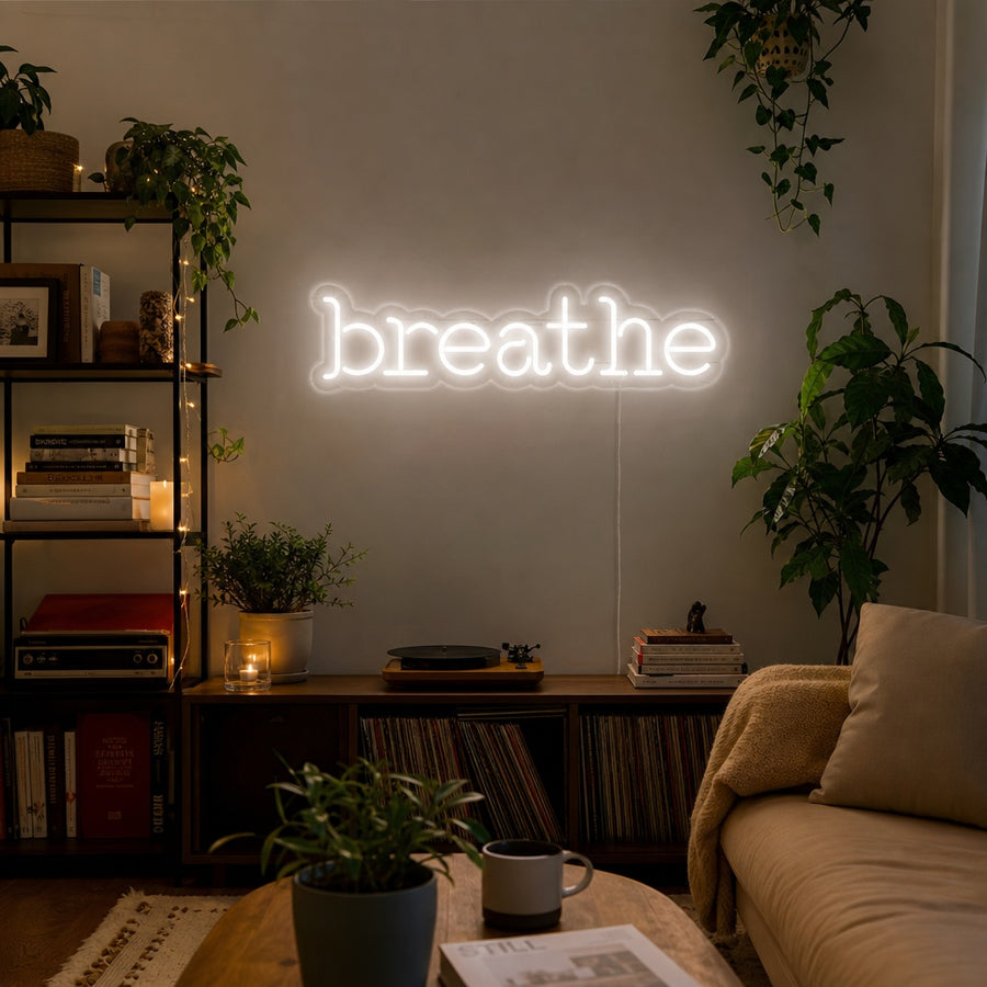 White 'Breathe' script neon sign glowing above a bed in a modern, serene bedroom, next to a shelf with plants and books.