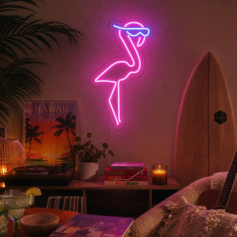 Vibrant pink flamingo neon sign glowing pink on a textured grey wall above a modern grey sofa in a brightly lit room with a potted plant.