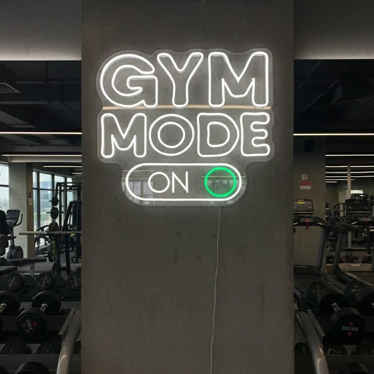 A "Gym Mode On" neon sign glowing powerfully on a dark wall inside a bustling fitness center, surrounded by exercise equipment and weights.