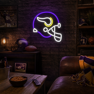 A lifestyle shot of the lit 'Minnesota Helmet' neon sign glowing brightly on a brick wall in a rustic home bar setting, surrounded by subtle sports memorabilia.