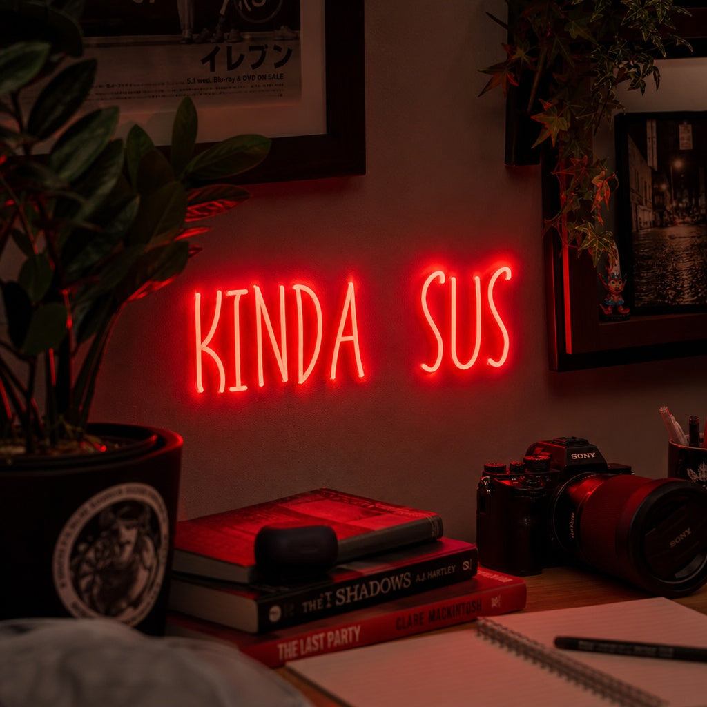 A 'KINDA SUS' neon sign glowing in a game room setting, mounted on a textured dark wall above a desk with gaming monitors and accessories.