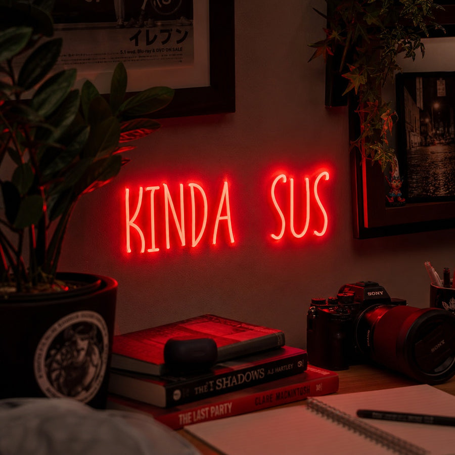 A 'KINDA SUS' neon sign glowing in a game room setting, mounted on a textured dark wall above a desk with gaming monitors and accessories.