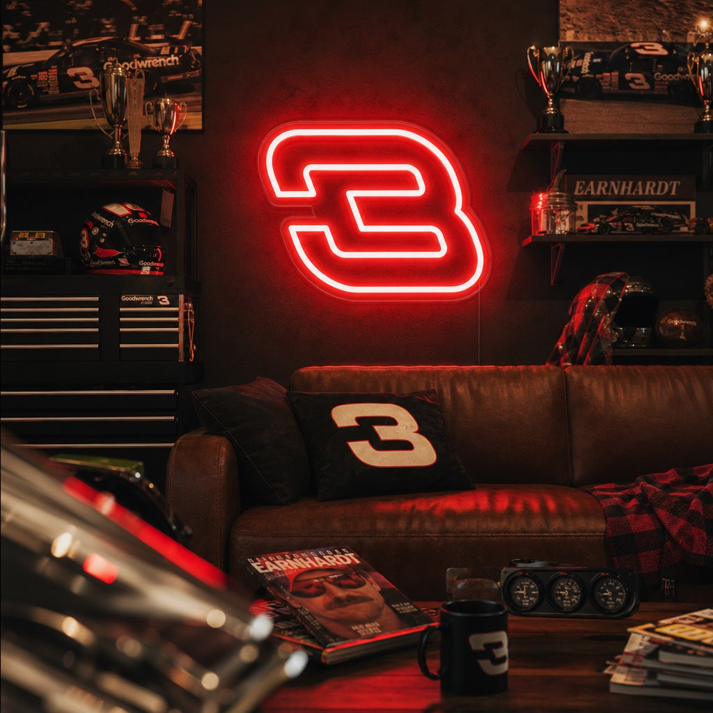 A glowing NASCAR No. 3 neon sign mounted on a textured brick wall in a rustic, well-lit garage, positioned above automotive tools and memorabilia.
