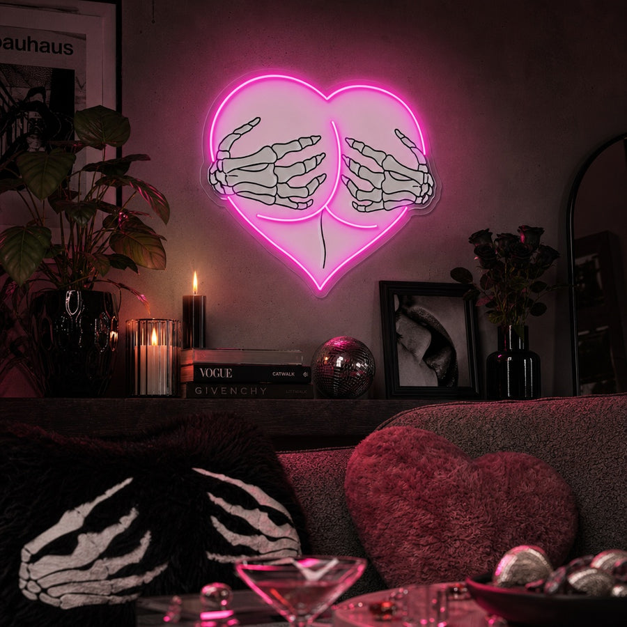 UV print neon sign depicting glowing pink skeleton hands framing a heart shape on a pair of hips, displayed against a blurred interior background.