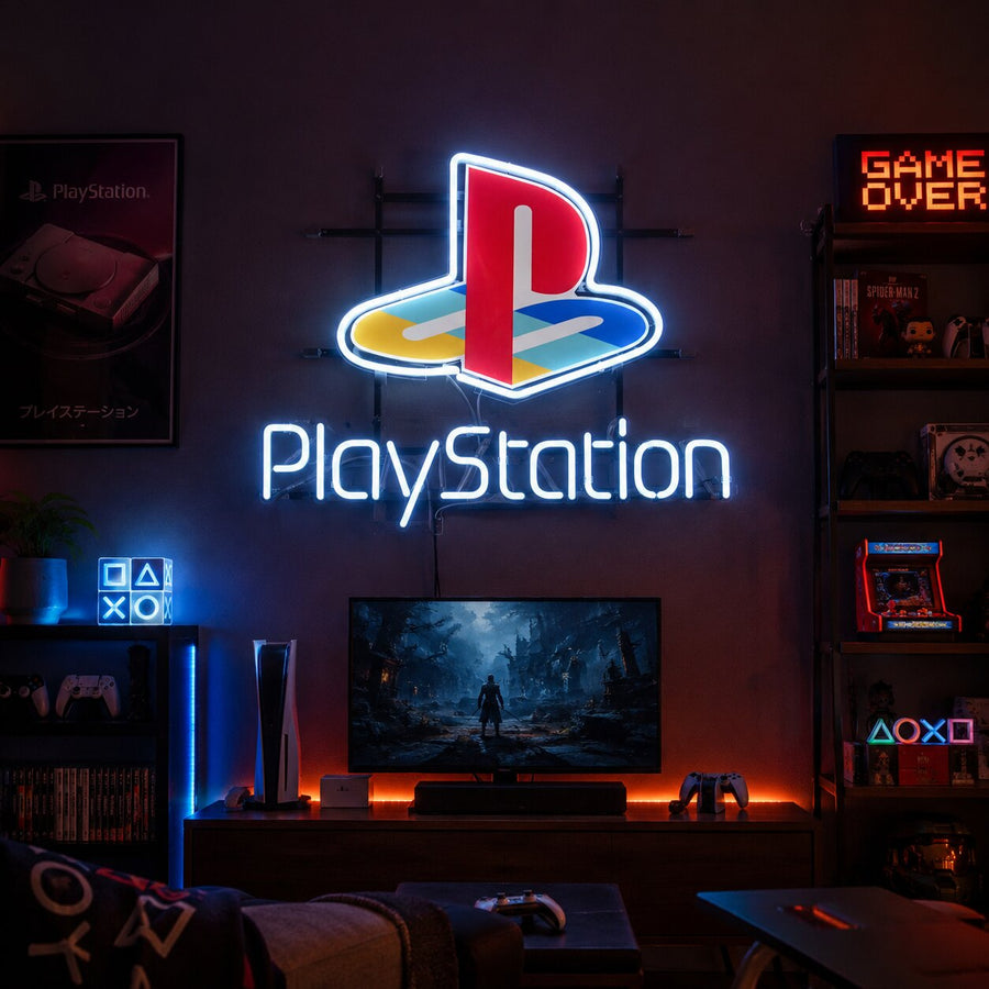 A vibrant 'Play Gaming Station' neon sign glowing in various colors (green triangle, red circle, blue X, pink square symbols, and white text) mounted on a wall. The image clearly shows the physical construction including the glass tubes, internal wiring, and the transformer/power unit.