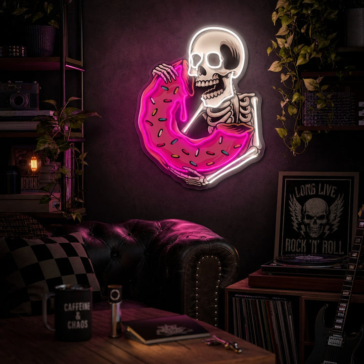 A vibrant UV print neon sign depicting a skull eating a colorful donut, glowing on an exposed brick wall above a cozy table setting with mugs and plants.