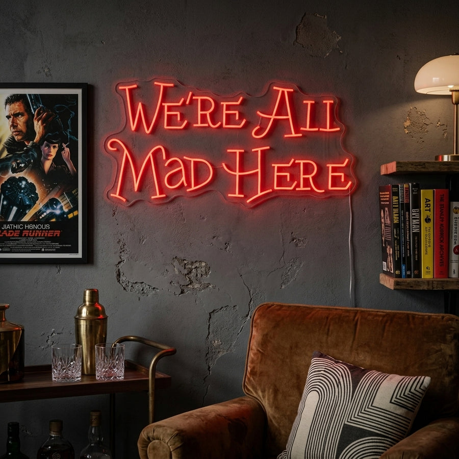 A glowing 'We're All Mad Here' neon sign, styled after the Cheshire Cat, hanging as wall decor above a sofa and plants in a cozy living room setting.