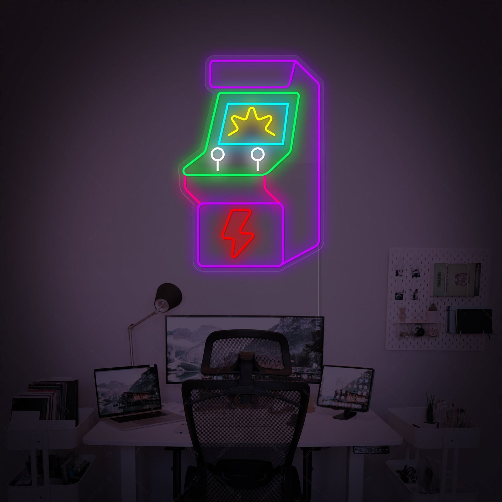 Glowing neon sign in the shape of a retro arcade machine, illuminated in blue and pink on a dark wall, serving as decor for a computer desk and gaming area.