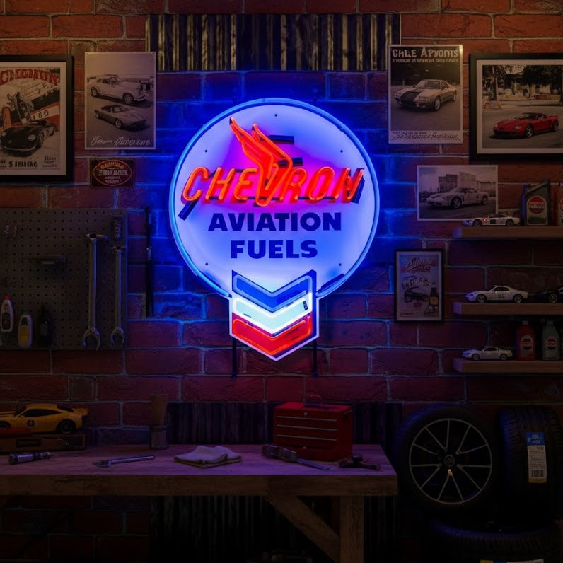A glowing retro Chevron Aviation Fuels neon sign featuring blue, red, and white glass tubes mounted on a brick wall in a vintage-style garage or workshop.