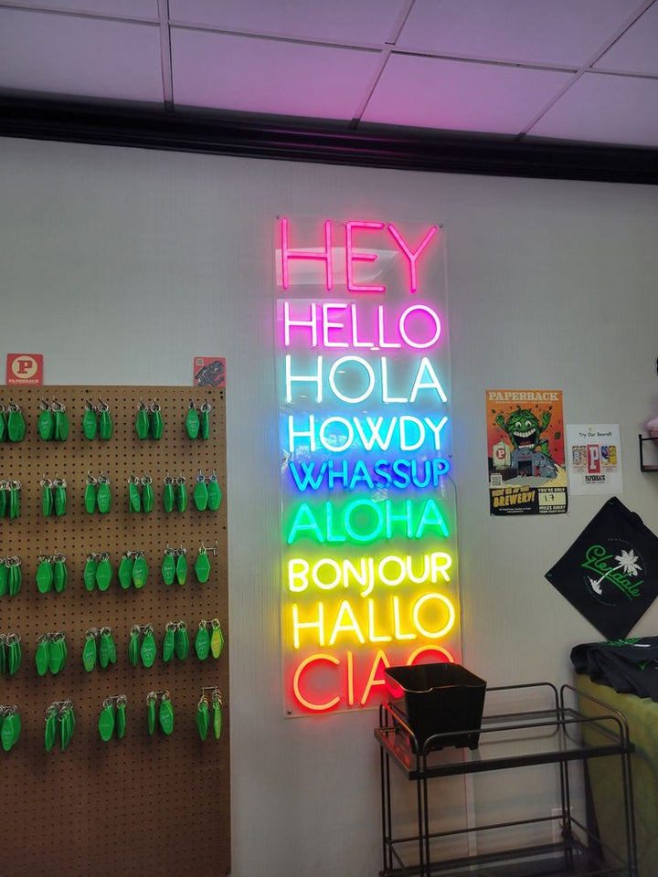 Multilingual hello neon sign spelling 'Hey Hello Hola Howdy Whassup Aloha Hallo Bonjour Ciao' with different colorful fonts on an acrylic backboard in a retail setting.