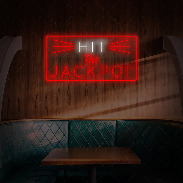 The glowing red neon sign displays 'HIT THE JACKPOT' in a bold, stylized font, mounted on a clear backboard and radiating light against a simple dark background.