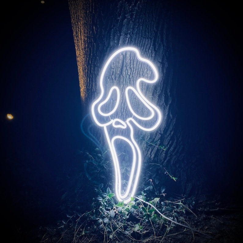 A bright, illuminated neon sign depicting the classic white Scream ghost face mask, casting an eerie white light against a dark, wooden background at night. The sign is hanging outdoors and is clearly detailed with neon tubing.