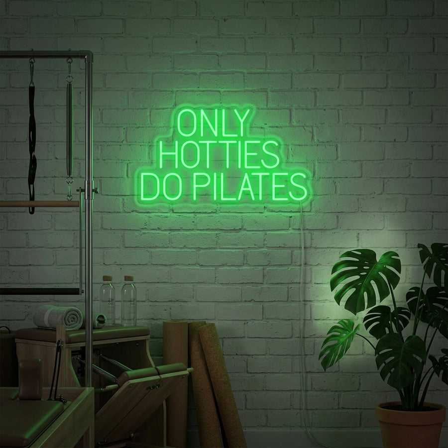 A vibrant 'Only Hotties Do Pilates' neon sign mounted on a textured brick wall in a well-lit Pilates studio with natural light and plants.