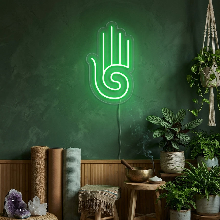 A glowing Hamsa Hand with a figure in a yoga pose inside, rendered in bright white neon tubing, mounted on a stone textured wall above a shelf with plants and a statue.