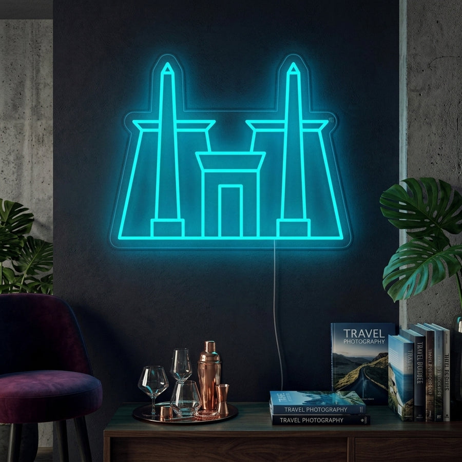 A lifestyle photograph of an illuminated orange neon sign depicting the ancient Egyptian Karnak Temple complex mounted on a modern dark blue living room wall above a grey sofa and potted plants.