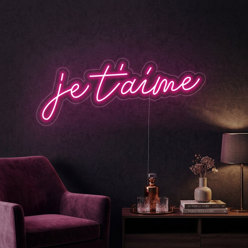 A lifestyle photo showing a flowing script 'Je T'aime' neon sign glowing with a warm pink light on a textured white wall, placed above a cozy arrangement with a wooden shelf and green plants.