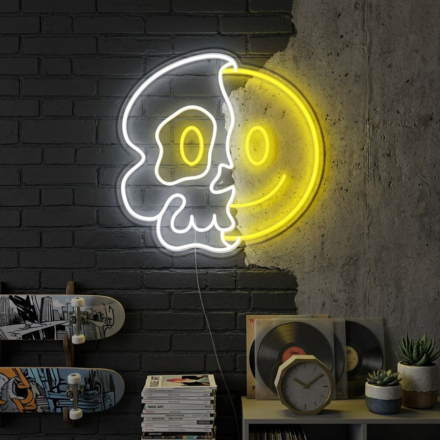A lifestyle photo of the illuminated 'Half of Skull with Smile Face' neon sign hanging above a rustic wooden desk with gaming peripherals and art prints, casting a vibrant glow in a moody, modern room.