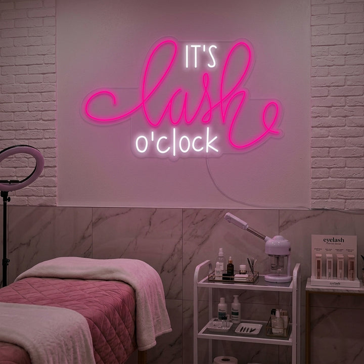 'Its Lashes Oclock' pink neon sign glowing brightly on a textured gray wall, creating a playful and glamorous atmosphere in a beauty studio setting.
