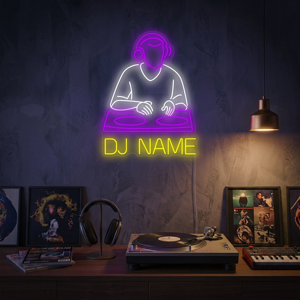A custom DJ logo neon sign in a vibrant pink glow, featuring 'DJ' in large block letters above 'YOUR LOGO HERE' in a script font, hanging on a dark textured wall next to a DJ deck and mixer setup in a dimly lit studio or club.