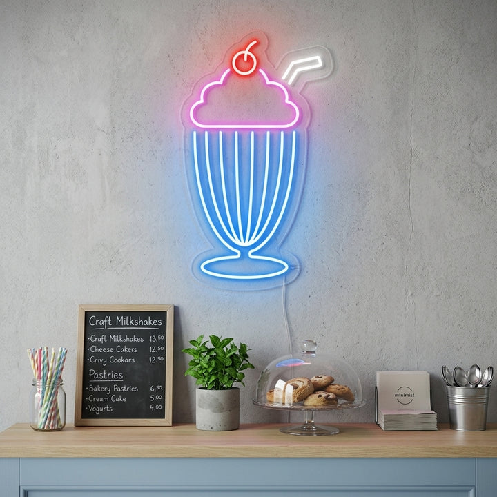 Glowing pink Milkshake neon wall sign hanging on a kitchen wall next to cafe-style shelving.