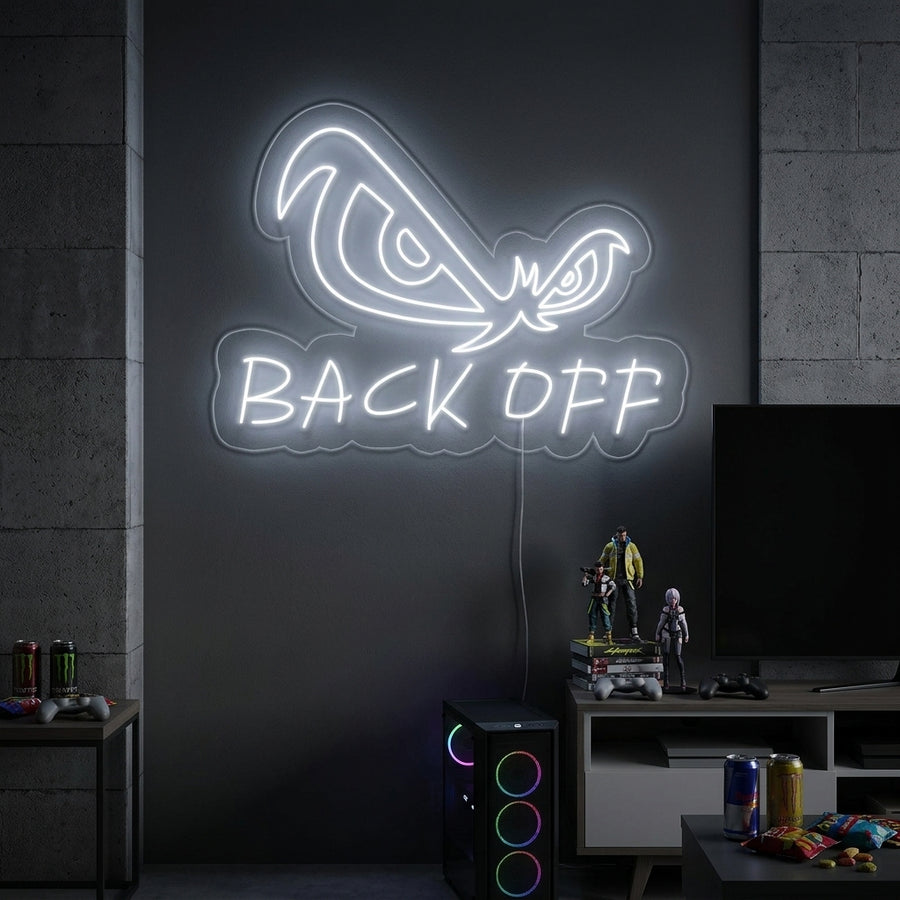 A vibrant 'Back Off Eagle Eyes' neon sign glows blue in a contemporary living room setting, mounted on a grey wall above a sleek black sofa with patterned cushions.