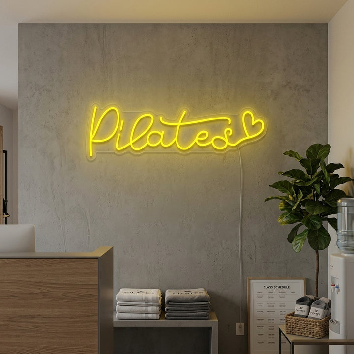 Lifestyle image showing a illuminated "Pilates Heart" neon sign glowing in a modern pilates or yoga studio setting with various mats and equipment visible.