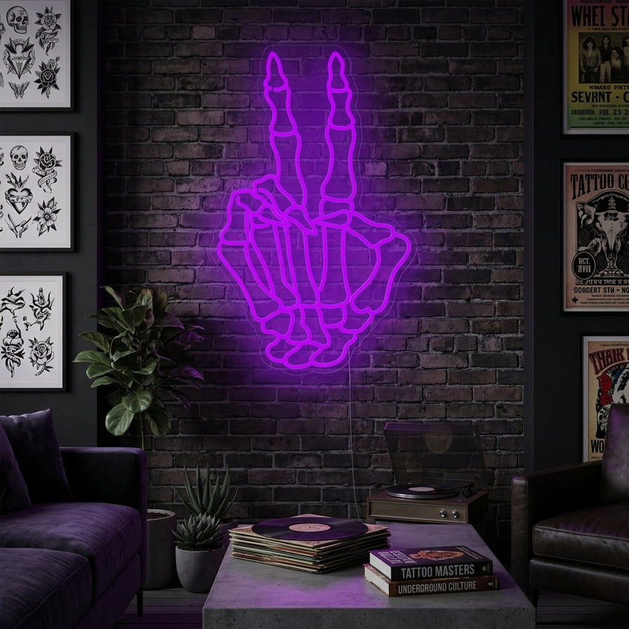 A close-up photograph of the lit-up Skeleton Hand Making Peace Sign Neon Sign, rendered in white light and hanging above a cozy bed with decorative pillows and bedding in a stylish room.