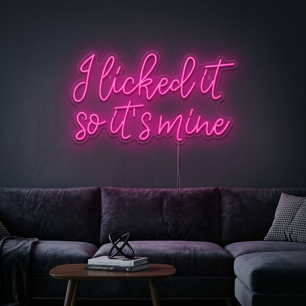 A neon wall sign reading 'I Licked It So Its Mine' in a stylized script, glowing warmly against a bedroom wall above some books and plants.