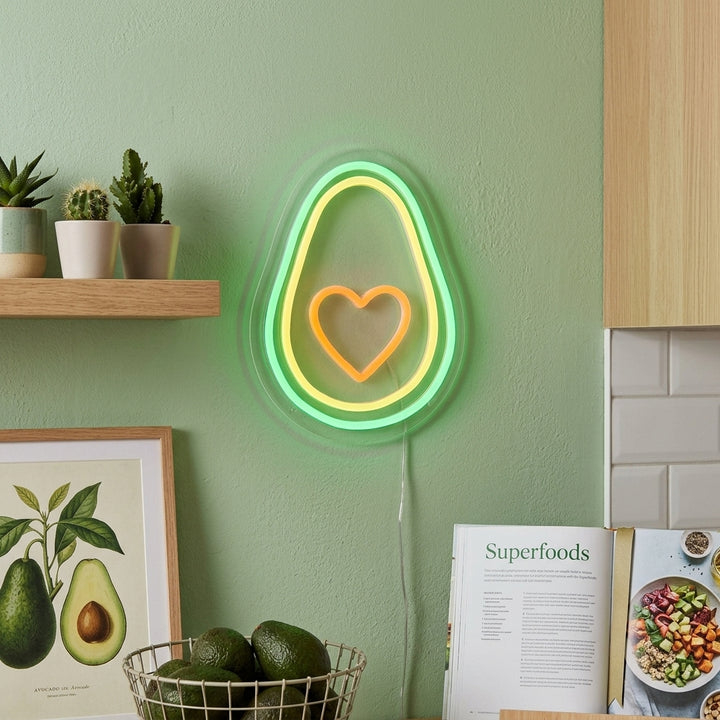An Avocado Heart Neon Sign glowing warmly above a stylized kitchen counter with fresh fruit and potted plants.