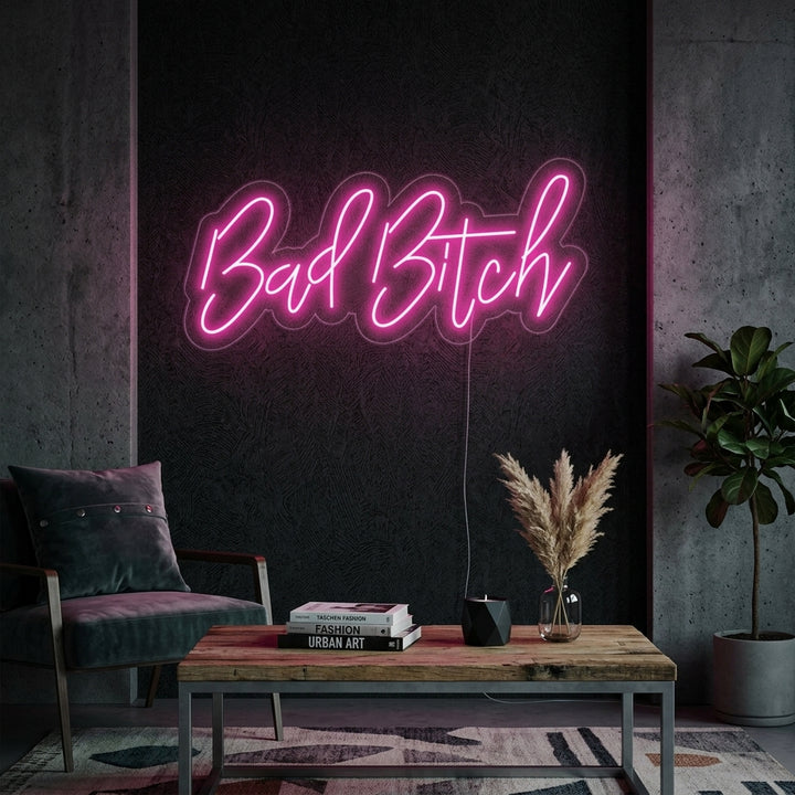 A vibrant 'Bad Bitch' script neon sign glowing with a warm light, mounted on a grey wall above a stylized, rustic-modern side table and lamp.