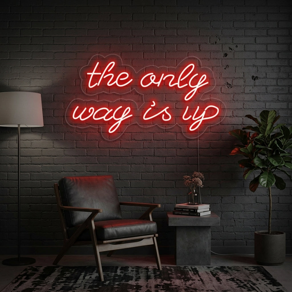 A lifestyle photograph showing the glowing white 'The Only Way Is Up' script neon sign mounted on a textured blue brick wall above a styled desk setup with a potted plant and books.
