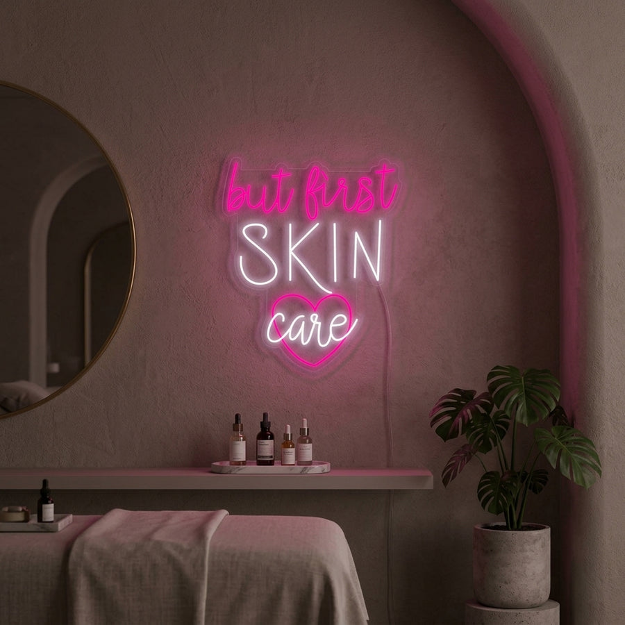 A glowing pink 'But First Skin Care' neon sign displayed on a white textured wall in a modern beauty setup, positioned above a vanity with skincare products and a mirror.