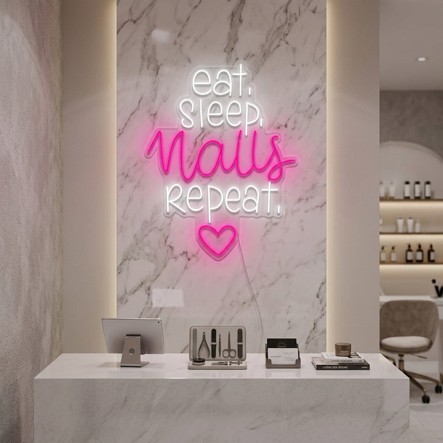 A close-up photograph capturing the glowing neon sign reading Eat Sleep Nails Repeat mounted on a dark textured wall in a modern salon setting. It features bright blue and pink lettering with glowing icons above a neat manicure station with polished bottles and tools.