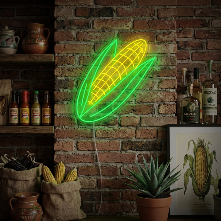 A lifestyle photo of the 'Corn' neon sign, glowing in warm yellow and green, mounted on a rustic wood plank wall above a kitchen counter filled with fresh corn and vegetables.
