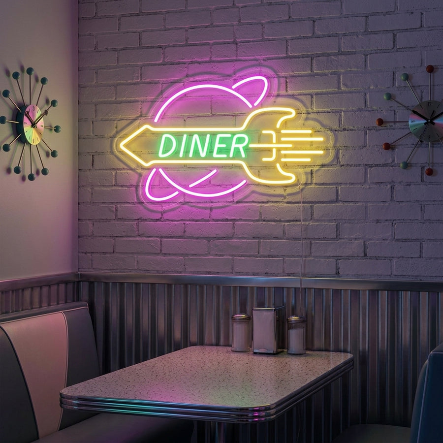 A vibrant ROCKET Rocket Diner neon sign showing a blue and red rocket over bold yellow and red script text, casting a colorful glow against a patterned diner wall above a booth.