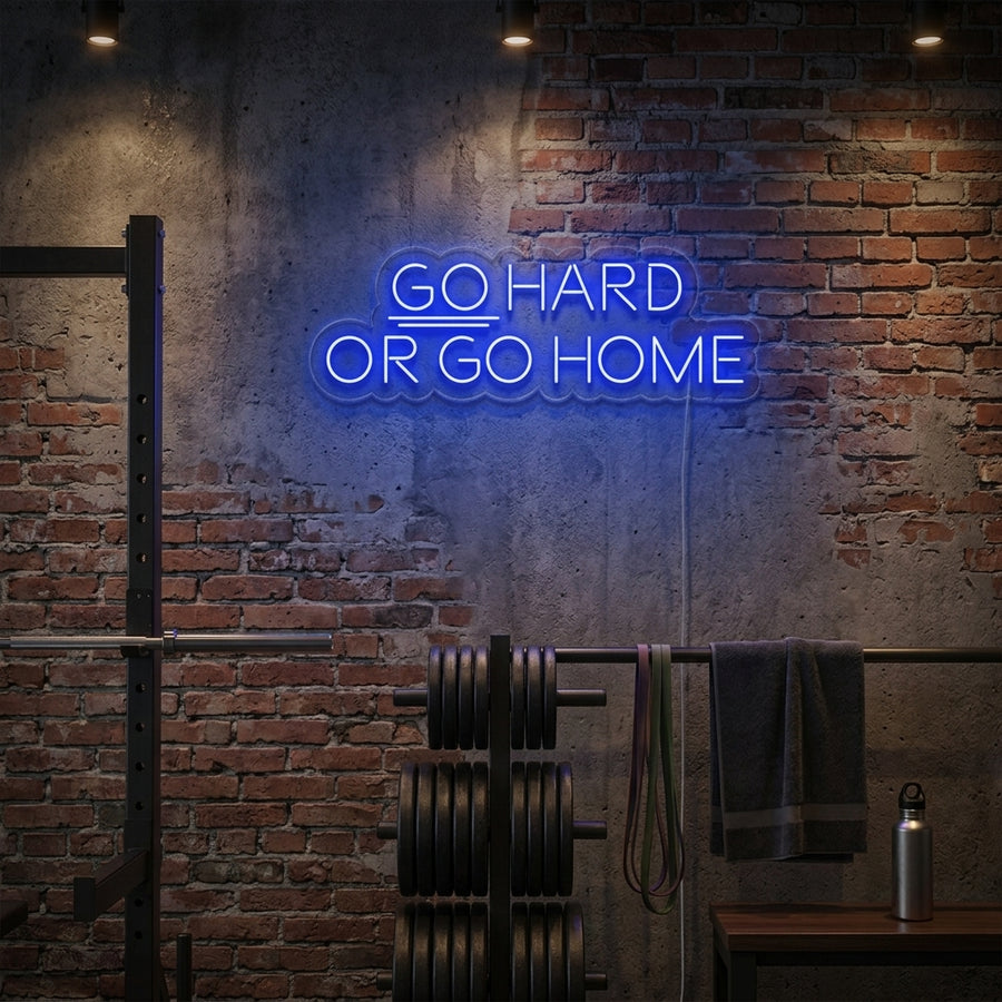 A lifestyle shot of the lit 'Go Hard or Go Home' neon sign mounted on a dark textured wall within a well-equipped home gym setting, casting an inspiring glow over a squat rack and weights.