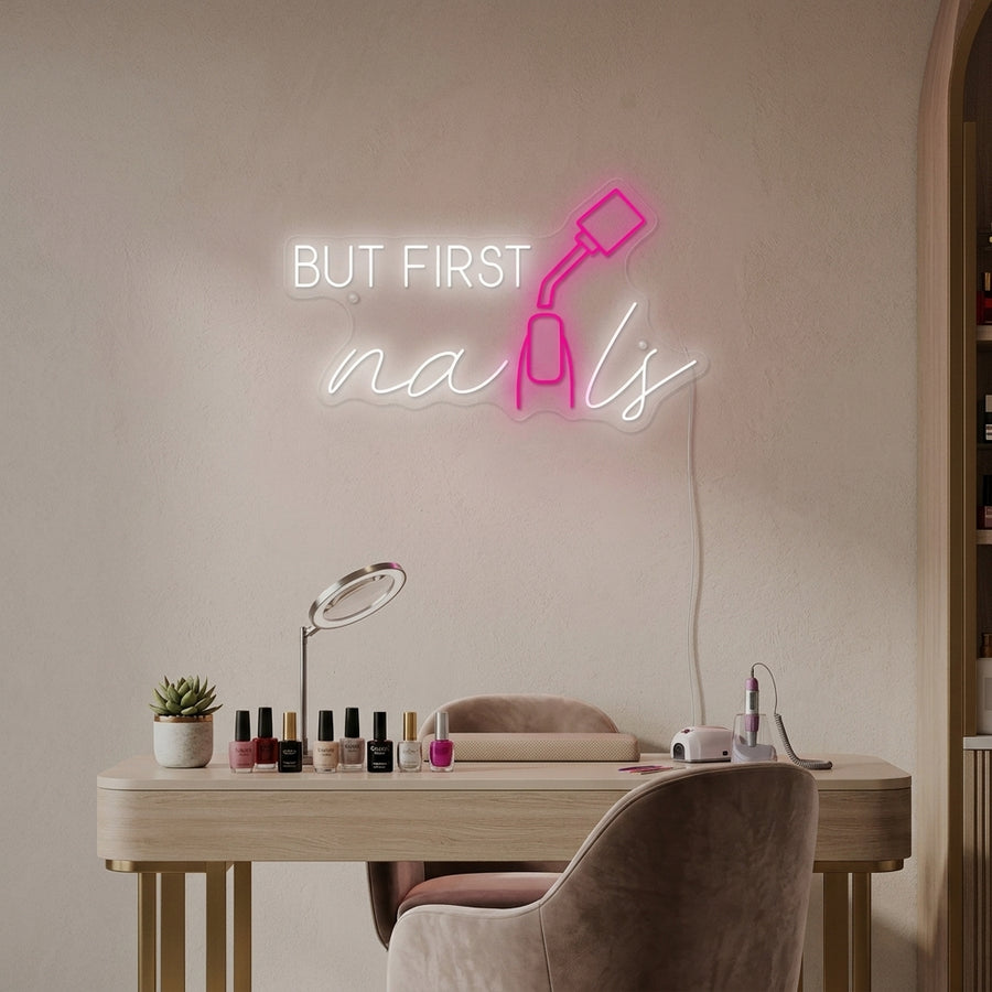 'But First Nails' pink neon sign displayed above a manicure station in a stylish nail salon setting.