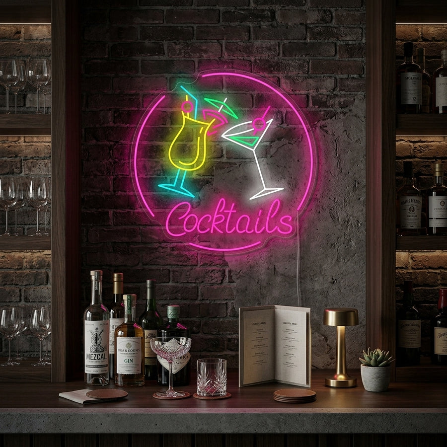 A glowing pink 'Cocktails' neon sign mounted on a dark brick wall in a rustic, modern bar setting, positioned above glass shelves stocked with liquor bottles and a stack of books.