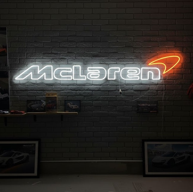 A close-up photograph showing a real 'Mclar-Car' style automotive neon sign glowing orange, mounted in a personal garage setting next to a shelf with framed photos.