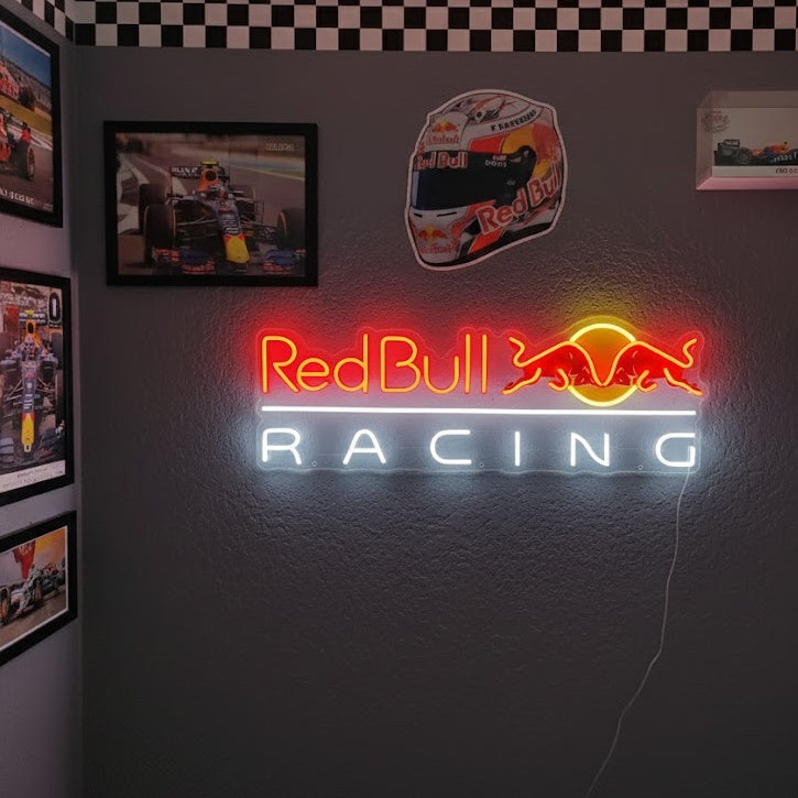A close-up photograph of a red and white illuminated Racing neon sign with a checkered flag accent, mounted on a dark textured wall within a garage filled with automotive memorabilia and a vintage car.
