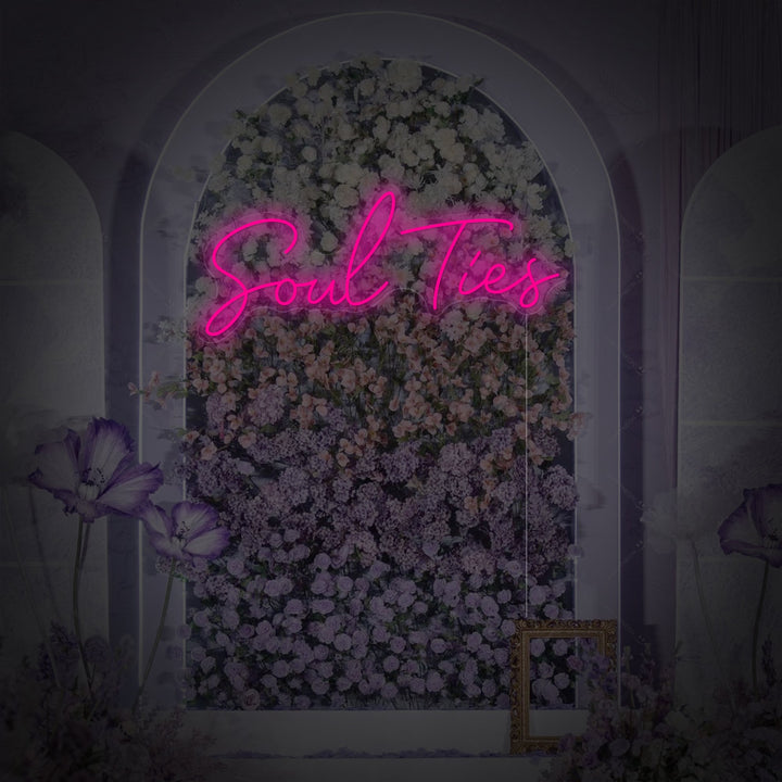Close-up photograph of the Soul Ties neon wall decor, showcasing the sleek acrylic backing, detailed script font, and the power cord. The sign is subtly glowing, mounted on a rustic wooden wall with soft, blurry decorative lighting in the background.