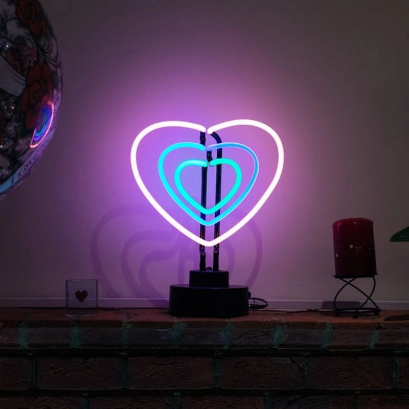 A table neon sign featuring three interconnected hearts glowing brightly in red, mounted on a dark base and displayed in a cozy living room setting on a brick ledge.